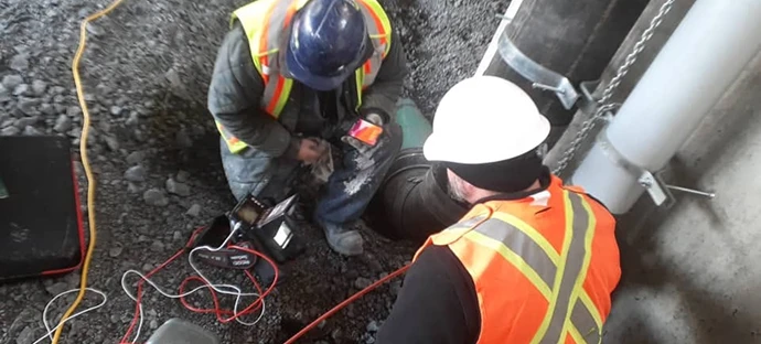 Two technicians wearing hard hats and safety vests inspecting an underground pipe using a camera system monitor.