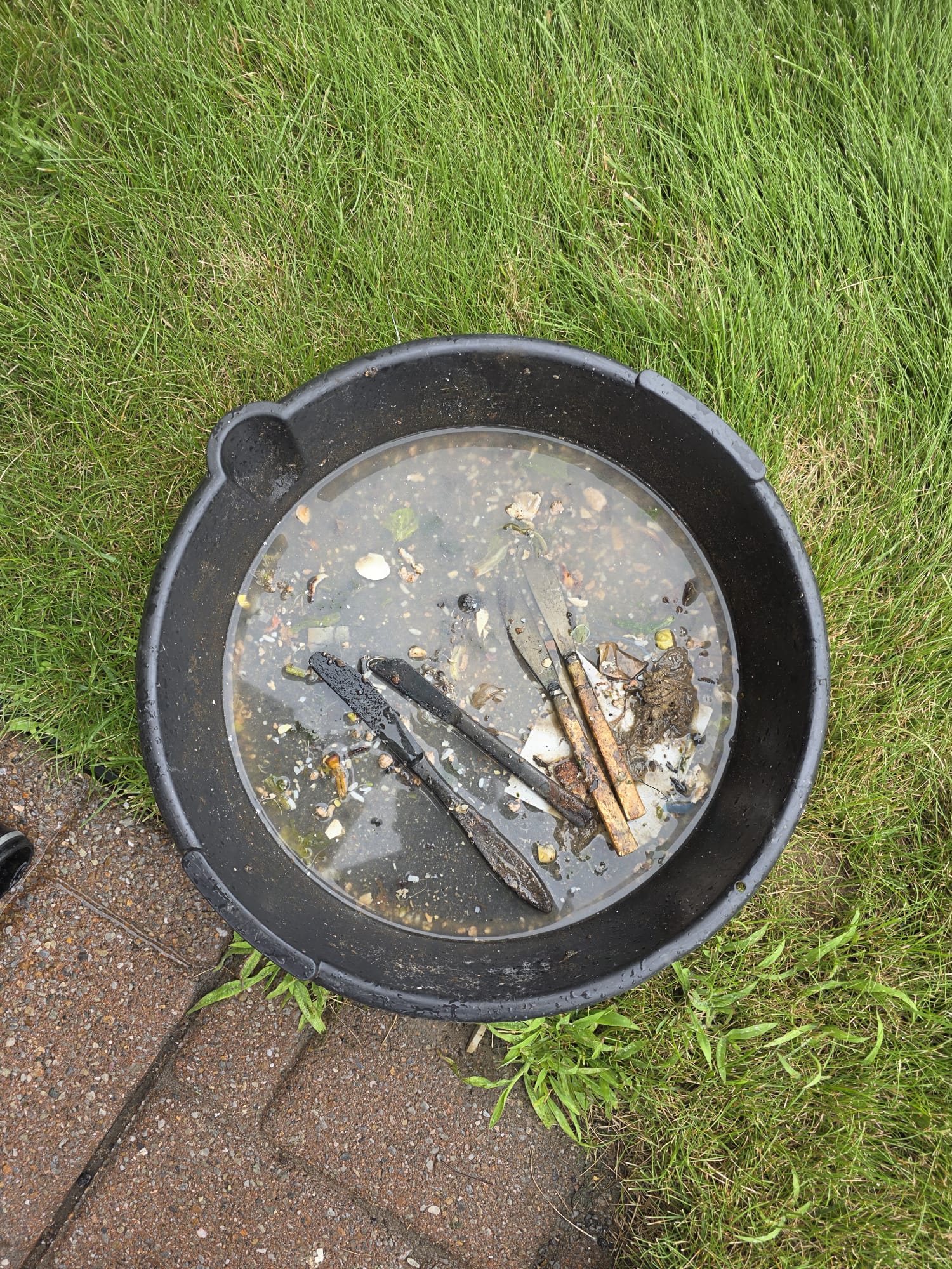 Bac de vidange contenant de l'eau, du gravier et des couteaux en métal récupérés d'un drain d'évier nettoyé par Flow Pro Drain.
